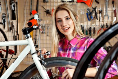 Aletlerin arka planında bisiklet atölyesindeki bir kadın. Pembe ekose gömlekli bir sarışın bir bisikleti tamir ediyor. Profesyonel bisiklet servisi kavramı.