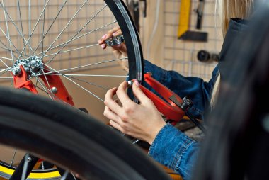 Aletlerin arka planında bisiklet atölyesindeki kadın. Mavi kot gömlekli sarışın bir bisikleti tamir eder. Profesyonel bisiklet servisi kavramı.