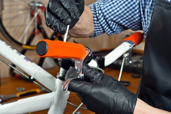 A man in a black apron repairs a bicycle handlebar. Master's hands close up. Repairman in gloves with tools.