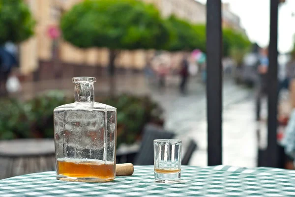 Bottle of alcohol near a glass on an empty table. The concept of alcoholism, loneliness and depression. Old vintage bottle on a checkered tablecloth. Blurred city street on background.