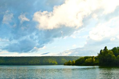 Göl ve orman manzarası. Bulutlu mavi gökyüzünün arka planında bir nehir. Gölün üzerinde gün batımı.