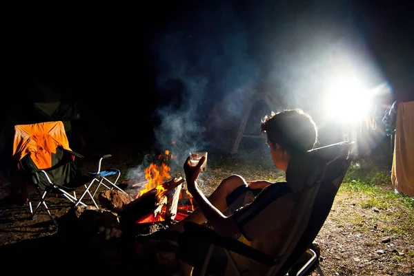 男が火事で椅子に座っている キャンペーンで夜の休息 火とテントを背景にアルコールのガラスを持つ男のシルエット 芸術的効果としての劇的な光との対比 — ストック写真