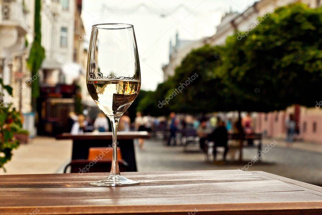 Glass of white wine close up. Alcoholic drink on a wooden table on a background of the city. Transparent wine on the summer terrace of the restaurant. Blurred people in the background.