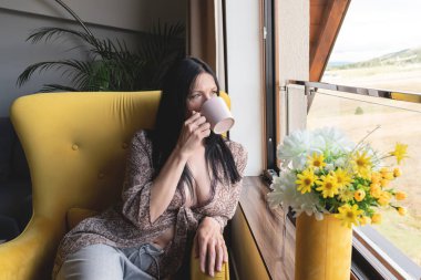 Güzel bir kadın kahve içiyor, koltukta oturuyor ve penceresinden manzarayı seyrediyor.