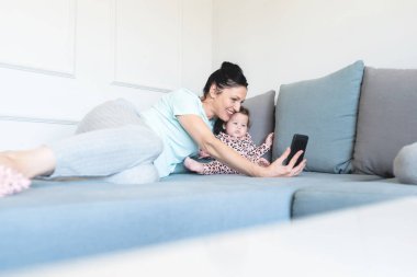 Sevgi dolu anne, sevimli kızıyla selfie çekiyor. Evdeki koltukta cep telefonu kamerası kullanıyor.