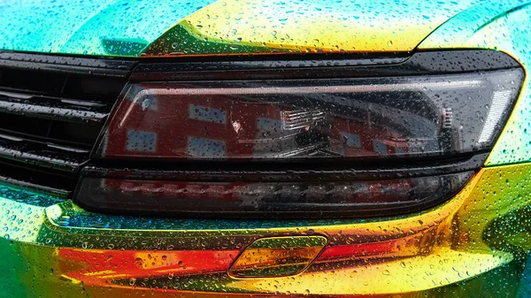 Chameleon holographic colour car in the rain. Car wrapping with iridescent paint. Raindrops on the car. Front view closeup. Modern LED headlights. Car wrapping. Wide photo web format.