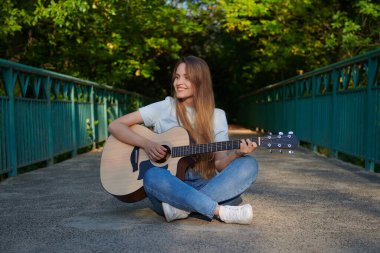 Portrait of brown-haired caucasian girl with acoustic guitar at the park. Beautiful sunset light on the bridge. Musician sitting on the bridge and playing guitar. Smiling teenager. Freedom concept.