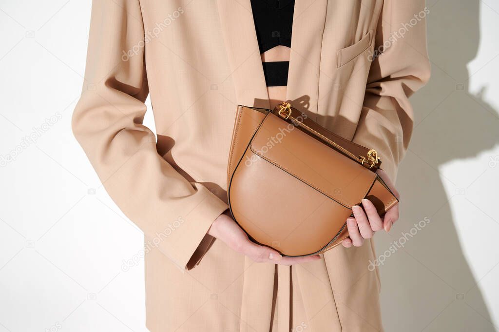 Brown leather handbag in girl's hand. Person with luxury bag in beige classic suit. Horizontal photo. Place for logo.