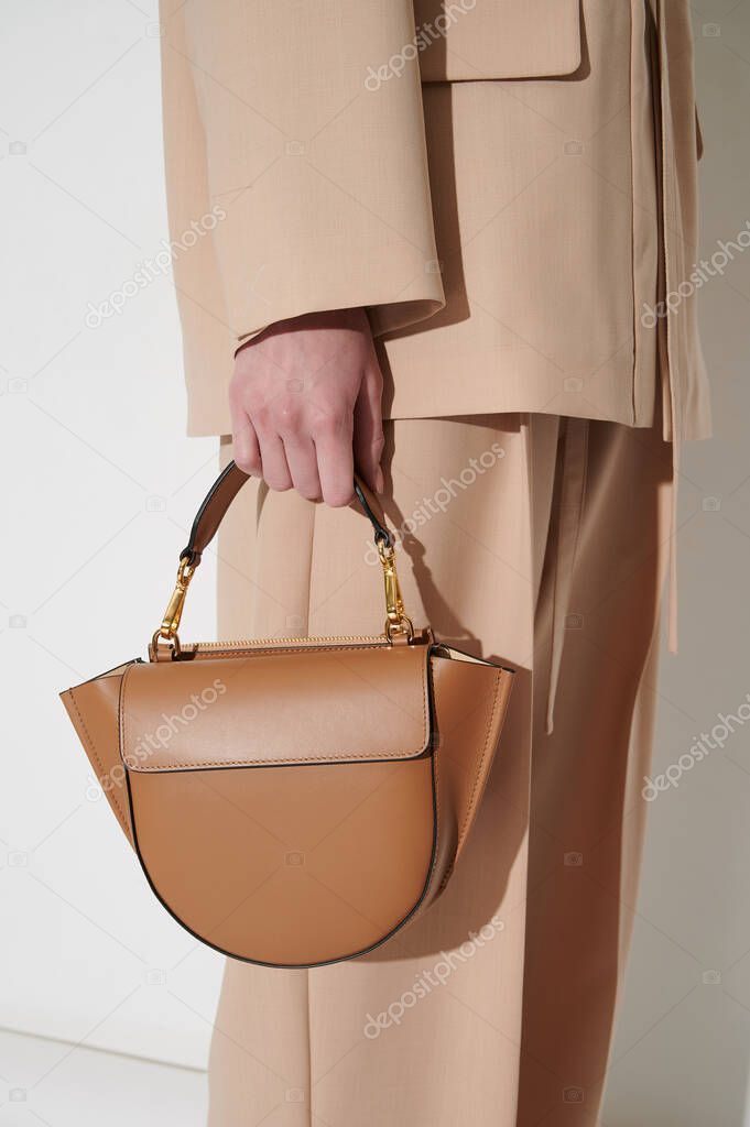 Brown leather handbag in girl's hand. Person with luxury bag in beige classic suit. Vertical photo.