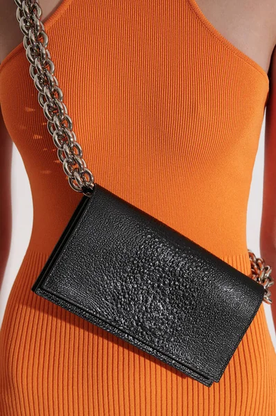 Girl in tight orange dress with black leather handbag. Luxury bag made from embossed leather with reptile effect with huge metal chain. Front view closeup.