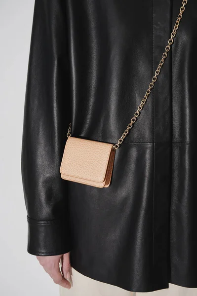 Beige leather embossed crossbody wallet on a golden chain. Person in black leather jacket. Small leather handbag. Cowhide leather. Vertical photo.