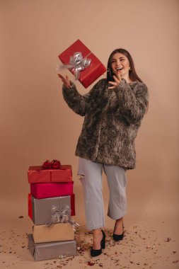 Yanında hediyeler olan kısa kürklü mutlu bir kız bej rengi arka planda izole edilmiş bir fiyonk hediye kutusu fırlatıyor.. 