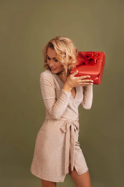 An interesting short haircut blonde girl in a shiny dress shaking the red gift box isolated over an olive color background.