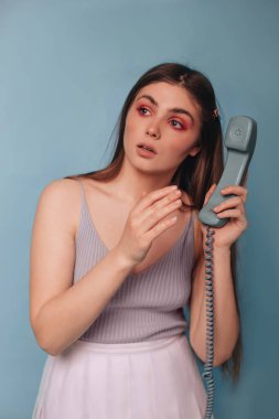 Parlak makyajlı, beyaz etekli bir kız mavi arka planda izole edilmiş bir telefon ahizesi tutuyor. Kız çağırıyor..