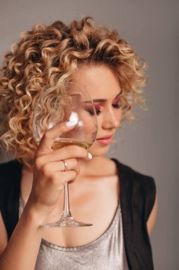 Afro kıvırcık saçlı ve profesyonel makyajlı mutlu bir kız bir şişe alkolle ayakta duruyor, gri arka planda bir bardağı ileri doğru tutuyor..