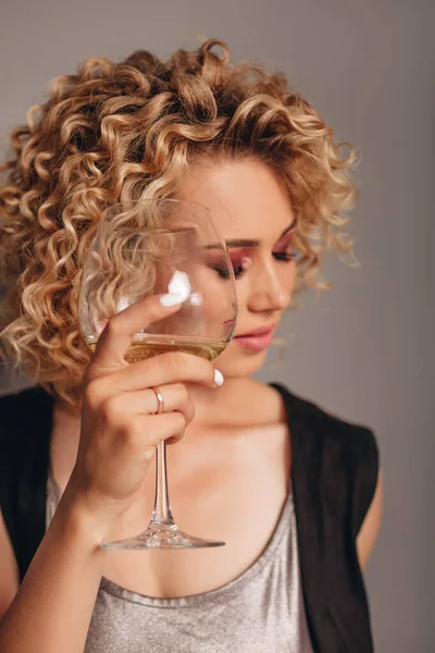 Afro kıvırcık saçlı ve profesyonel makyajlı mutlu bir kız bir şişe alkolle ayakta duruyor, gri arka planda bir bardağı ileri doğru tutuyor..