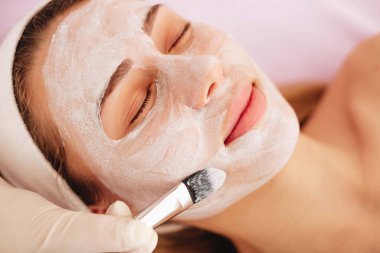 Deri maskeli genç bir kadının yüzü. Güzellik uzmanı, müşterinin yüzüne fırça yardımıyla beyaz bir maske takıyor.. 