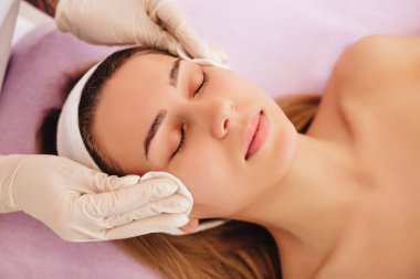 Beyaz eldivenli güzellik uzmanı pamuklu pedlerle genç bir kadının yüzünü siliyor. Pamuk süngerlerle yüz temizleme. Hijyen. Profesyonel cilt bakımı.