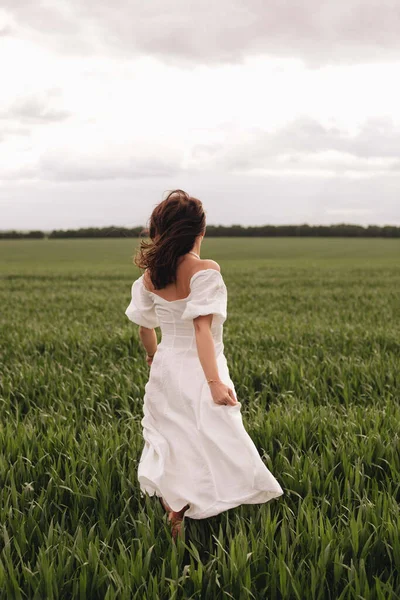 A beautiful brunette girl in a white linen dress runs along a field of green wheat. Stylish girl in the field from the back.