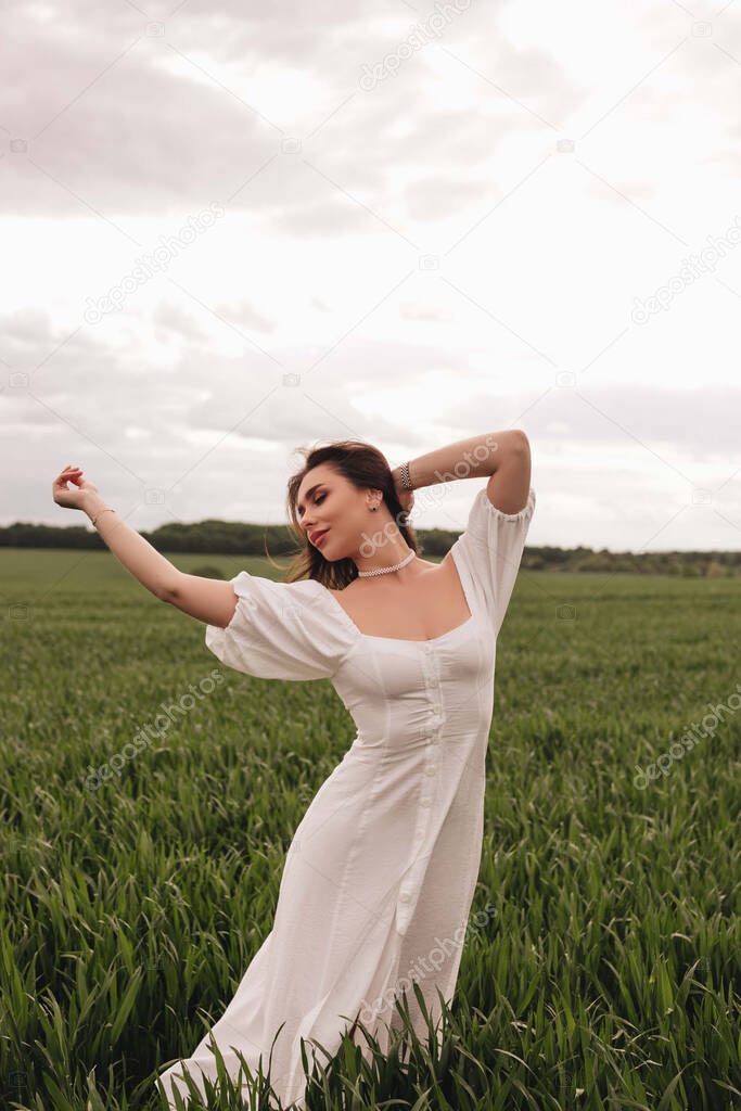 A beautiful brunette girl in a white linen dress is dancing in a green field of wheat. Stylish girl in the field.