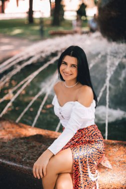 Şık giysiler içinde gülümseyen ve neşeli bir kız şehir merkezinde bir çeşmenin yanında dinleniyor. Yaşam tarzı kavramı. 