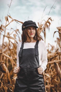 Beyaz kazaklı, siyah tulumlu ve bereli şık bir kız sonbaharda sarı mısır tarlasında duruyor..