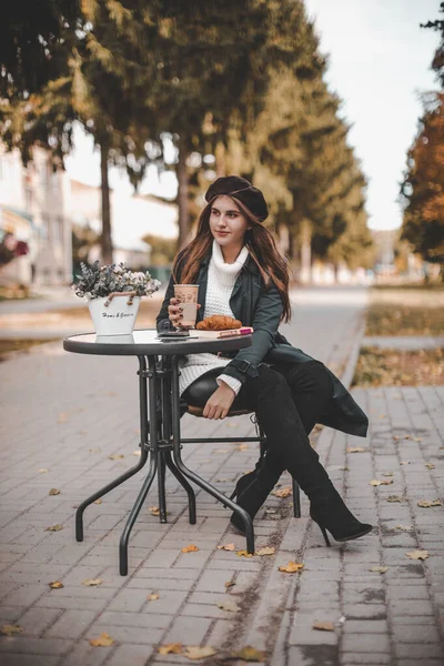 Yağmurluk ve siyah bereli güzel bir kız sonbaharda parktaki bir ara sokakta bir kafede kahve içiyor. Fransız bir kadın sonbahar parkında bir kafede kruvasanla kahve içiyor..