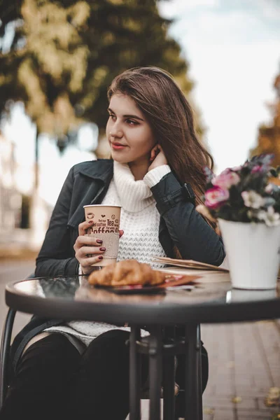 A beautiful girl in a raincoat and a black beret is drinking coffee at a table in a cafe on an alley in the park in autumn. A French woman drinks coffee with a croissant in a cafe in an autumn park.