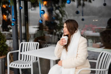 Beyaz kışlık ceketli ve eşarplı gülümseyen genç bir kadın açık bir kafede oturmuş kahvesini yudumluyor. Kar ve mavi Noel süsleri sıcak bir festival ortamı yaratır..