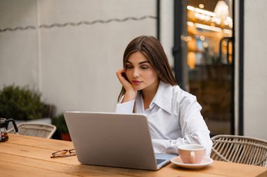 Açık kafede dizüstü bilgisayar kullanan, internette çalışırken kahve içen genç bir kadın. Sakin, modern atmosferli uzak iş, öğrenim ya da yaratıcı serbest çalışma konsepti.