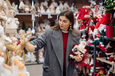 Noel süsleri ve mevsimlik süslerle dolu bir bayram mağazasında alışveriş yaparken iki Noel heykelciğini karşılaştıran genç bir kadın..