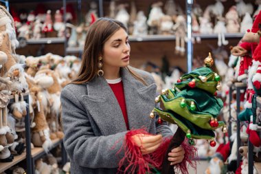 Noel ağacı oyuncağını inceleyen genç bir kadın bayram heykelcikleri ve mevsimlik süslemelerle dolu bir mağazada.