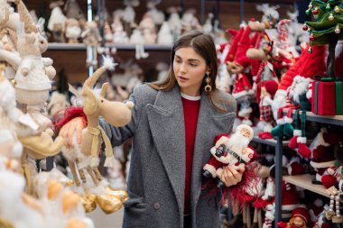 Noel süsleri ve mevsimlik süslerle dolu bir bayram mağazasında alışveriş yaparken iki Noel heykelciğini karşılaştıran genç bir kadın..