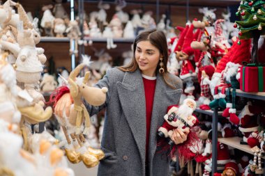 Noel süsleri ve mevsimlik süslerle dolu bir bayram mağazasında alışveriş yaparken iki Noel heykelciğini karşılaştıran genç bir kadın..
