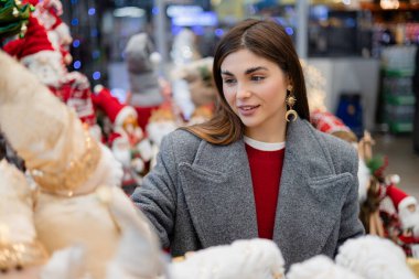 Noel oyuncakları ve şenlikli dekorasyonları seçen genç bir kadın mevsimlik süslerle çevrili bir tatil mağazasında..