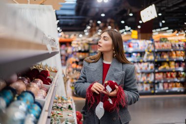 Noel süsleri ve şenlikli takılarla dolu bir mağaza koridorunda kırmızı Noel süsü seçen genç bir kadın..