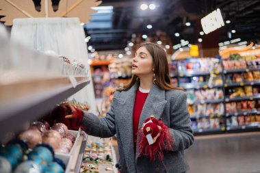 Noel süsleri ve şenlikli takılarla dolu bir mağaza koridorunda kırmızı Noel süsü seçen genç bir kadın..