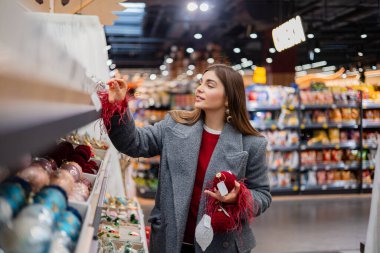 Noel süsleri ve şenlikli takılarla dolu bir mağaza koridorunda kırmızı Noel süsü seçen genç bir kadın..
