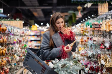 Genç bir kadın, bayram dekoruyla dolu bir alışveriş sepetini elinde tutarak, bir mağaza koridorunda Noel süsü bulurken şaşırdı..