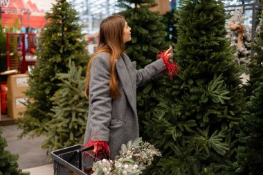 Noel ağacı seçen genç bir kadın elinde şenlikli süslemelerle dolu bir alışveriş sepeti tutuyor..