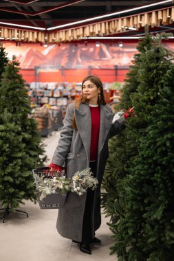 Noel ağacı seçen genç bir kadın elinde şenlikli süslemelerle dolu bir alışveriş sepeti tutuyor..