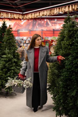 Noel ağacı seçen genç bir kadın elinde şenlikli süslemelerle dolu bir alışveriş sepeti tutuyor..