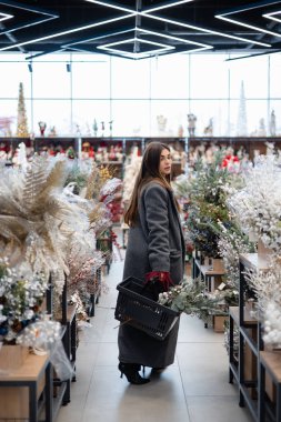 Elinde alışveriş sepeti tutan ve Noel yeşilliği ve şenlik düzenlemeleriyle dolu bir mağazada kış tatili süslemelerini seçen bir kadın..