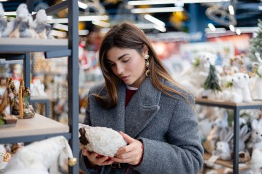 Bir tatil mağazasında Noel heykelciklerini inceleyen, elinde dekoratif bir melek tutan ve raflardaki mevsimlik dekorasyonlara bakan genç bir kadın..