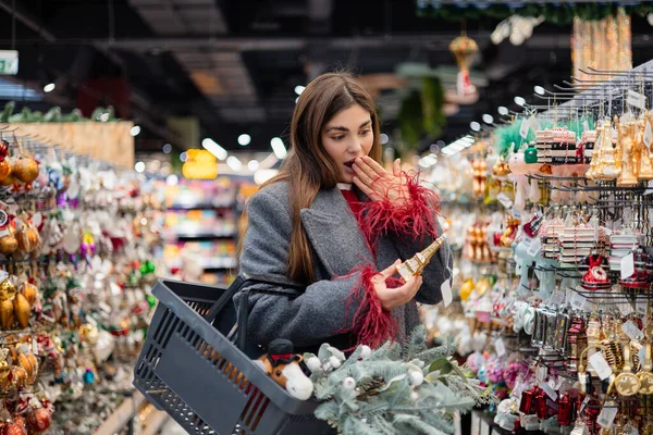 Genç bir kadın, bayram dekoruyla dolu bir alışveriş sepetini elinde tutarak, bir mağaza koridorunda Noel süsü bulurken şaşırdı..