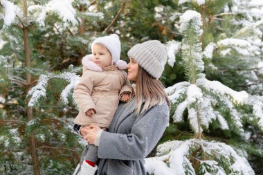 Anne kızını karlı bir çam ormanında tutuyor. Yumuşak ışık ve doğal atmosferli sakin bir kış anı..