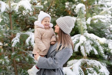 Anne gülen bebeğini karlı bir çam ormanında tutuyor. Sıcak kış anı neşe ve bağlarla dolu.