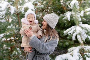 Anne gülen bebeğini karlı bir çam ormanında tutuyor. Sıcak kış anı neşe ve bağlarla dolu.