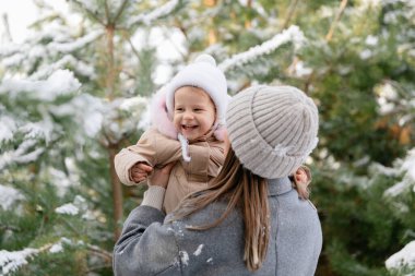 Anne gülen bebeğini karlı bir çam ormanında kaldırıyor. Doğal ışık ve kalın giysilerle neşeli aile kış anı.
