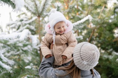 Anne gülen bebeğini karlı bir çam ormanında kaldırıyor. Doğal ışık ve kalın giysilerle neşeli aile kış anı.